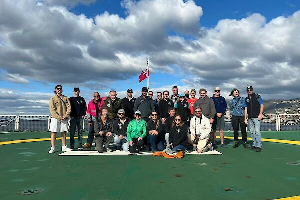 A group of smiling people standing in a group on a green helicopter deck on the back of a ship.