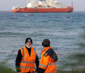 Two men in orange jackets stand talking on the beach with an orange ship in the ocean in the background