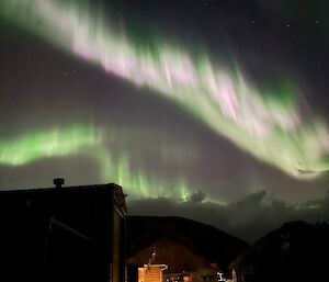 A pink and green aurora in the night sky above lit-up buildings