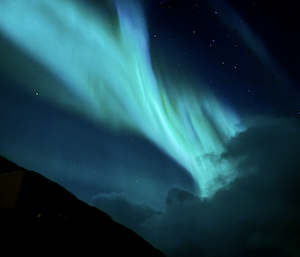 A vivid green-blue aurora reaches into the night sky with a dark hill and clouds in the foreground.