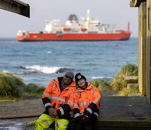 A smiling man and woman wearing bright orange coats sit on a step with an orange ship in the background.