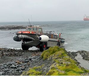 An amphibious boat with sharks teeth painted on the front leaves the sea and drives onto the land. An orange ship is in the background.