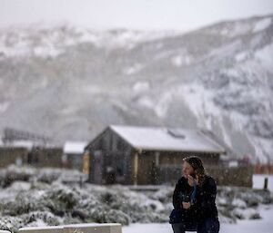 A women crouches in the snow and makes a radio call - a building and snow-covered mountain are in the background.