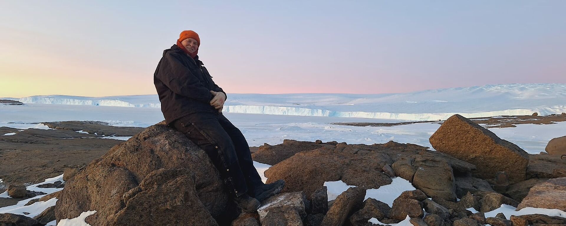A man sitting on a rock with a vast stretch of ice behind him and a soft pink glow from a sunset.