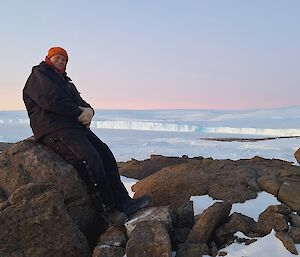 A man sitting on a rock with a vast stretch of ice behind him and a soft pink glow from a sunset.