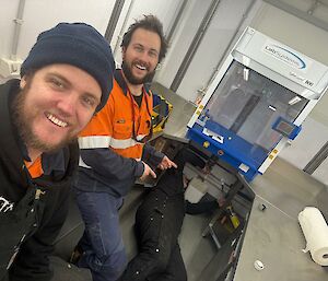 Two men smiling at the camera, while a third man lies on the floor fixing a pipe underneath some scientific equipment