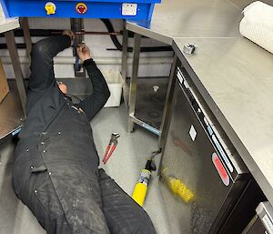 A man in black overalls lying underneath a sink trying to fix a pipe