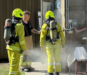 Two people wearing yellow firefighting uniforms about to enter a smoke-filled building.