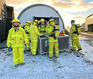 A group of people wearing new yellow firefighting outfits stand around a cage pallet collecting equipment.