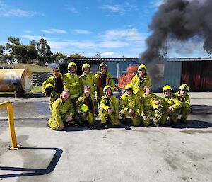 A group of smiling people wearing yellow firefighting outfits poses in front of a burning car.