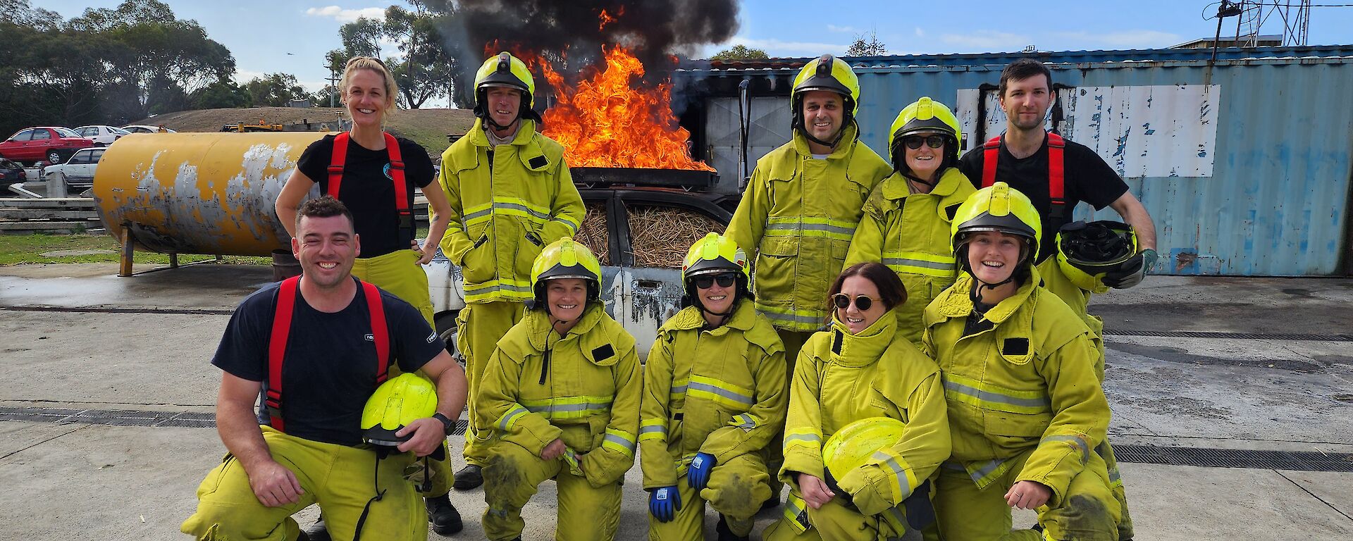 A group of smiling people wearing yellow firefighting outfits poses in front of a burning car.