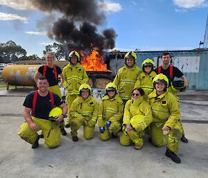 A group of smiling people wearing yellow firefighting outfits poses in front of a burning car.