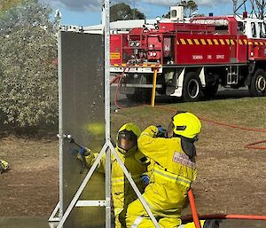 Two people in yellow firefighting outfits, kneel outside a door with an orange fire hose at the ready