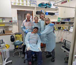 A smiling group of five standing in a doctor's surgery wearing surgical gowns
