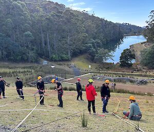 A group of people wearing helmets standing on a grassy hill practising the use of rescue ropes.