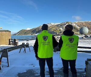 Two people wearing fluorescent yellow vests with Macquarie Island emblazoned on them facing a snow-covered landscape