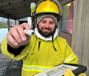 A smiling man wearing a firefighting outfit holds a clipboard and a texta, which he is pointing at the photographer.