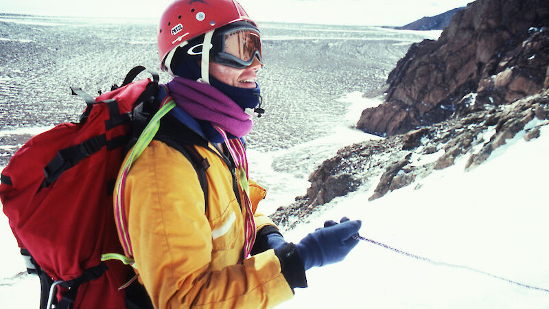 A man is standing holding a rope on a mountain in Antarctica. He is wearing a red helmet and googles. He has a yellow coat on and a red backpack.