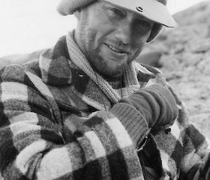 A black and white image of a man sitting down in a field. He has a peaked hat on and a checkered jacket.