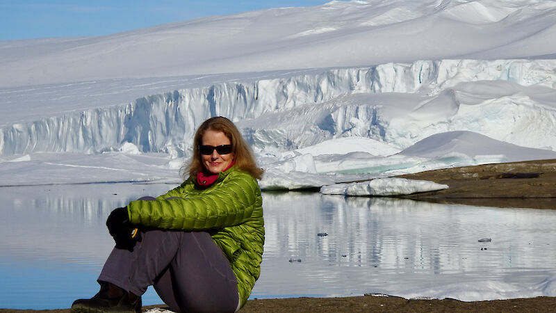 A woman sitting in front of a glacier in a green jacket.