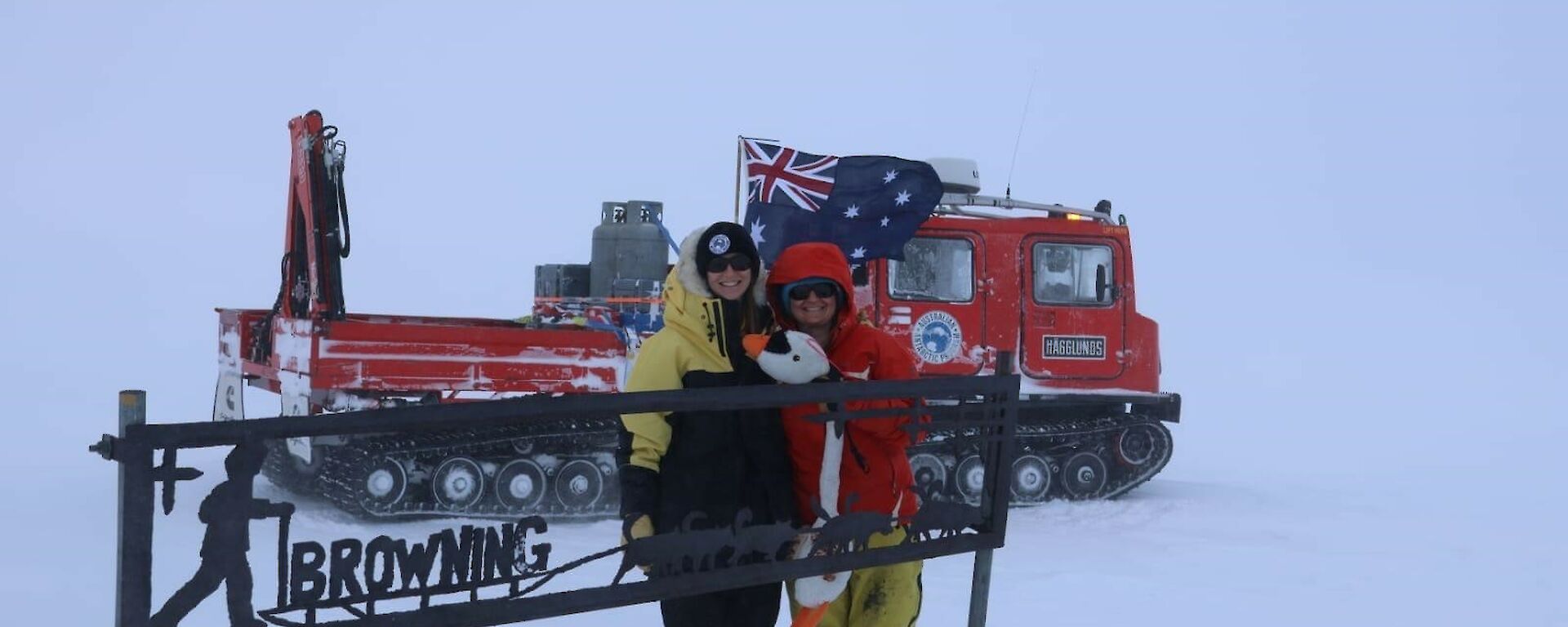 Two expeditioners and the goose standing in front of a sign in the snow fields
