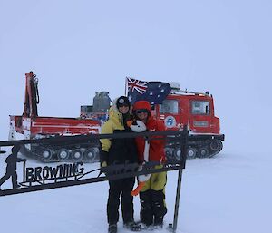 Two expeditioners and the goose standing in front of a sign in the snow fields