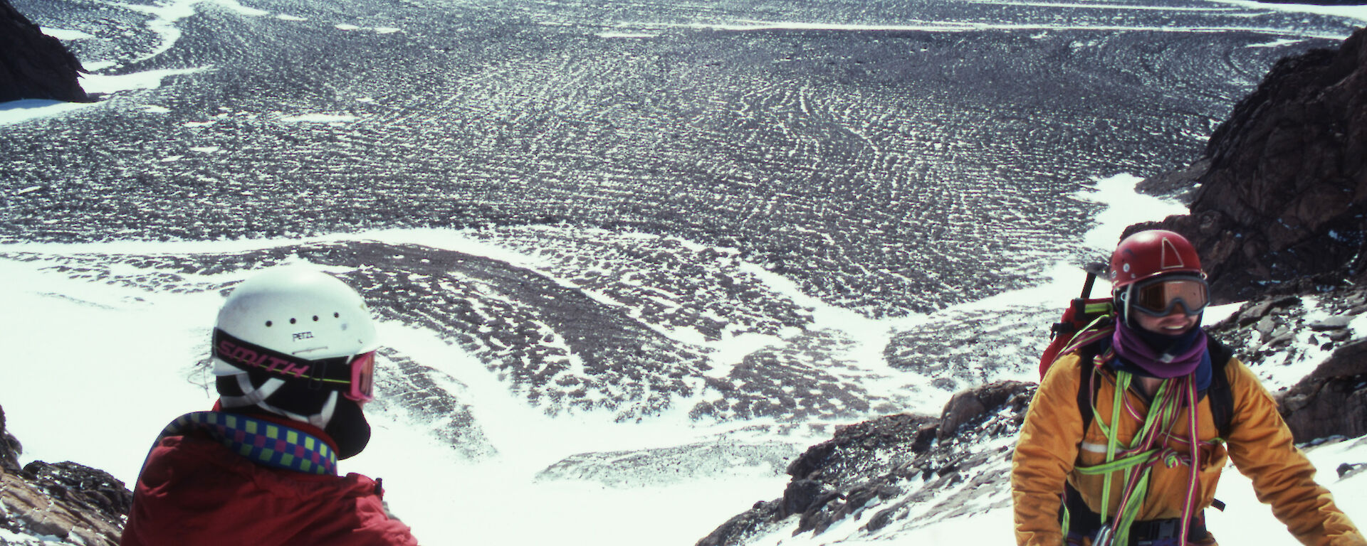 Two men wearing climbing equipment and helmets, kneeling in snow on a ridge high above a rocky landscape in Antarctica.