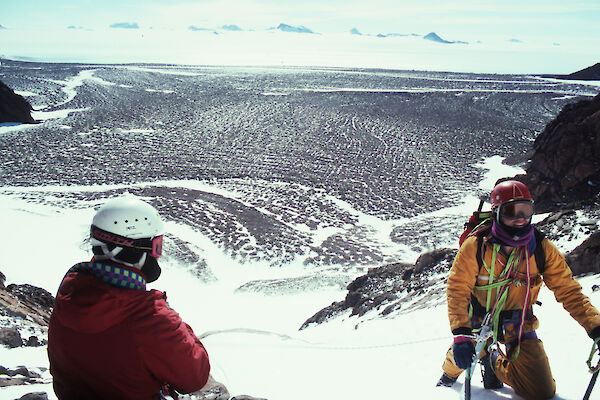 Two men wearing climbing equipment and helmets, kneeling in snow on a ridge high above a rocky landscape in Antarctica.