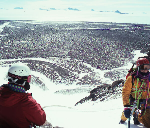 Two men wearing climbing equipment and helmets, kneeling in snow on a ridge high above a rocky landscape in Antarctica.