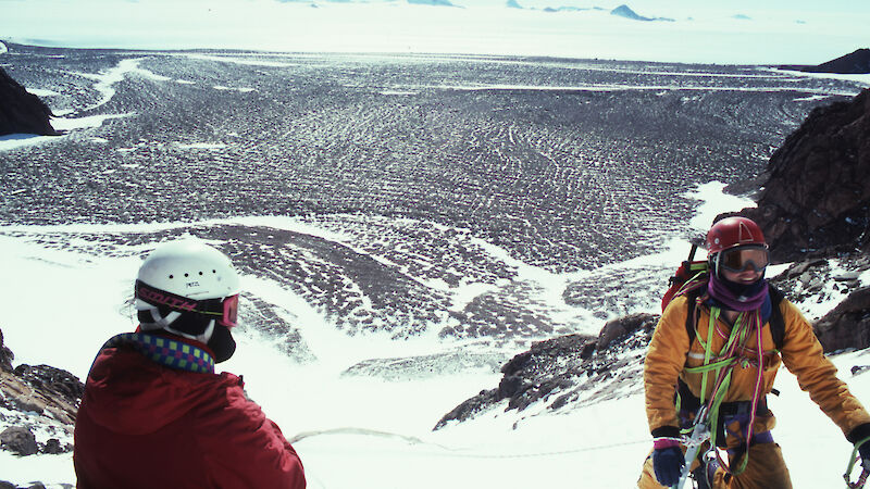 Two men wearing climbing equipment and helmets, kneeling in snow on a ridge high above a rocky landscape in Antarctica.