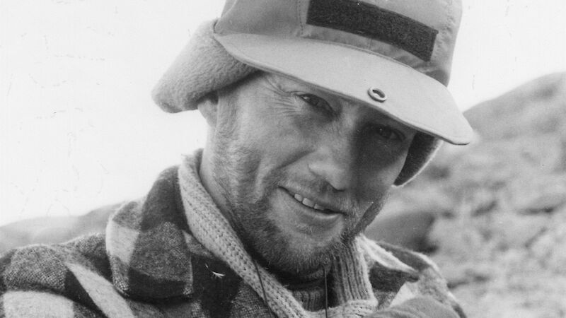 A black and white photo of a man wearing a checked woolen shirt and a fleece hat, sitting on a rocky hillside with a field notebook on his lap.