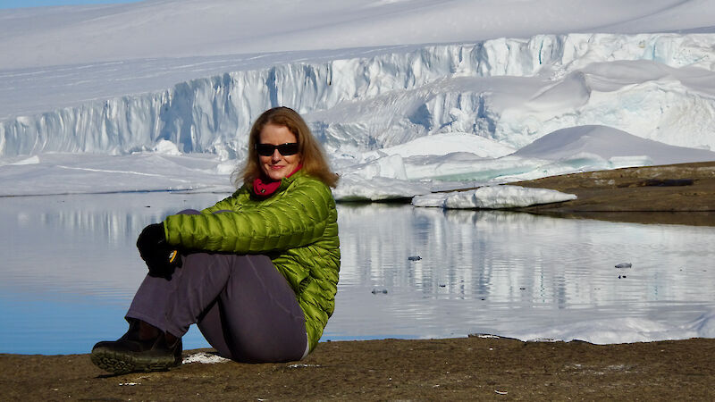 A woman in a green jacket and blue pants sits on rocks beside a bay, with an ice cliff rising in the background behind her.