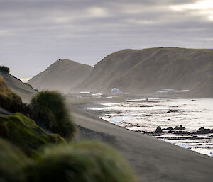 A hill covered in green tussock in the foreground, ocean to the right and two rugged green hills in the distance with buildings at the foot of the hills.