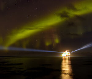 A green aurora dances over a ship in the water. The ship is well lit and has a blue strobe light emanating from either side into the dark night sky.