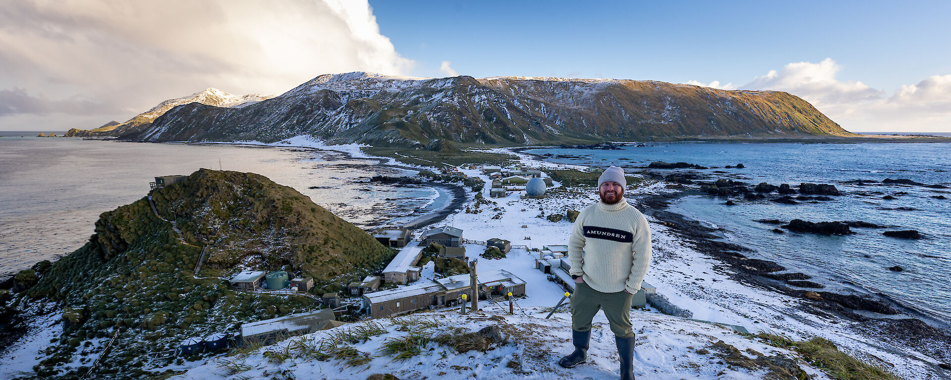 A man wearing a beanie and jumper stands on a snow-covered hill with a snow covered isthmus and plateau in the background.