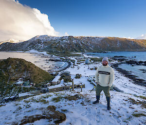 A man wearing a beanie and jumper stands on a snow-covered hill with a snow covered isthmus and plateau in the background.