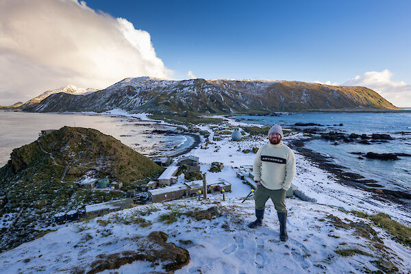 A man wearing a beanie and jumper stands on a snow-covered hill with a snow covered isthmus and plateau in the background.