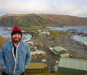 A smiling man in a beanie and denim jacket stands on a hill wiht an isthmus and plateau in the background.