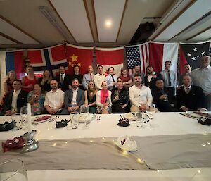 A group of well-dressed, smiling people behind a set dinner table with flags of different nations behind them.