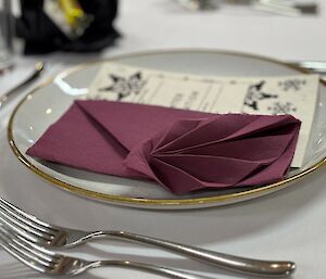 A gold rimmed plate with a menu and purple folded napkin. Silver forks in the foreground.