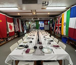 A long dining table set with plates, knives, forks and glassware with fancy folded napkins and dinner invitations. The walls of the dining room are adorned with flags from different nations.