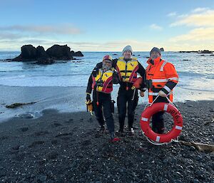 Two men in black dry suits and red and yellow life jackets stand next to a man in a bright orange jacket who is holding a life-ring. The backdrop is the beach.