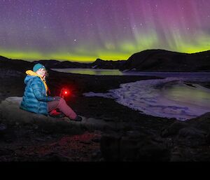 person sitting next to waters edge with aurora in sky