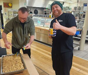 Two men stand in an cafeteria with a cake