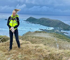 A smiling woman wearing outdoor gear stands atop a grassy hill with land and ocean behind and below her.