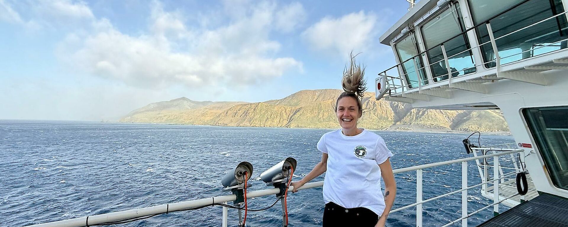 A smiling woman whose hair is swept upwards in the wind stands on the deck of a ship with an island visible in the distance