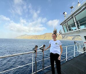 A smiling woman whose hair is swept upwards in the wind stands on the deck of a ship with an island visible in the distance