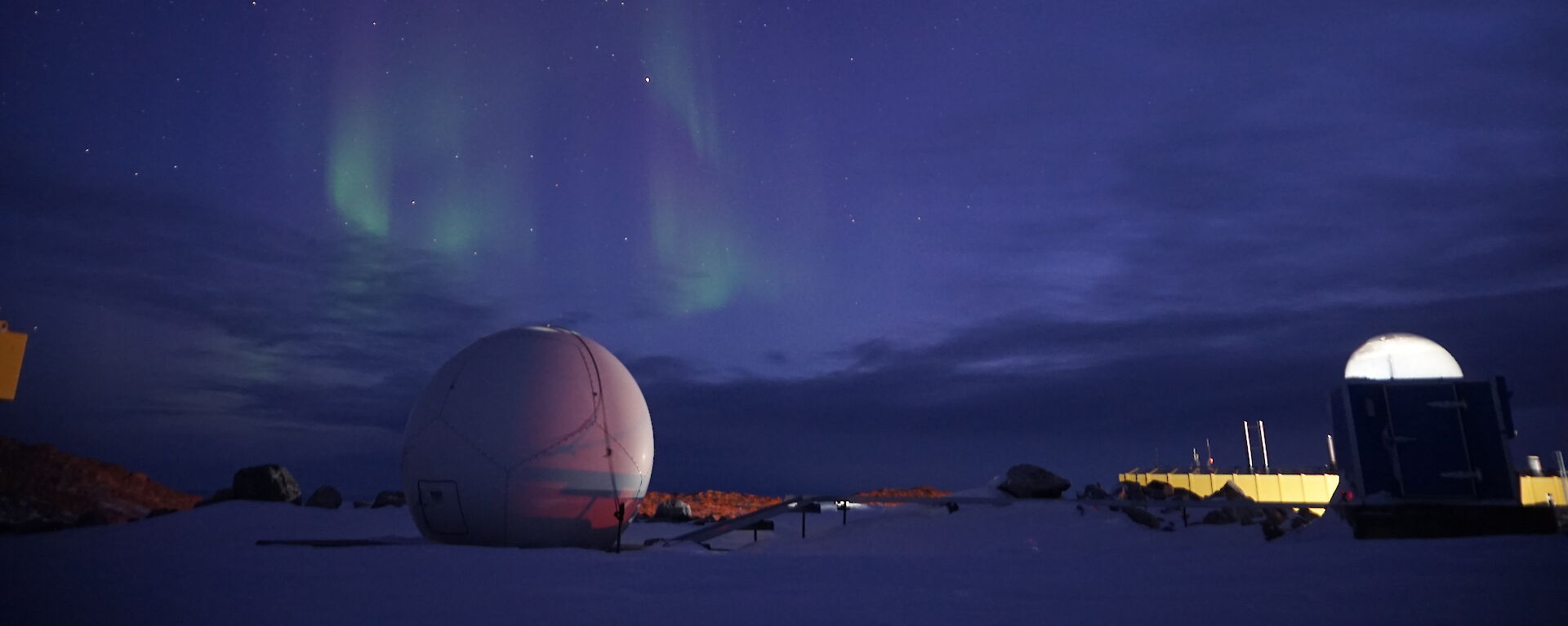Shimmering green light in a dark blue sky over station buildings and the satellite dome