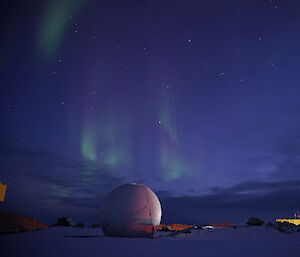 Shimmering green light in a dark blue sky over station buildings and the satellite dome