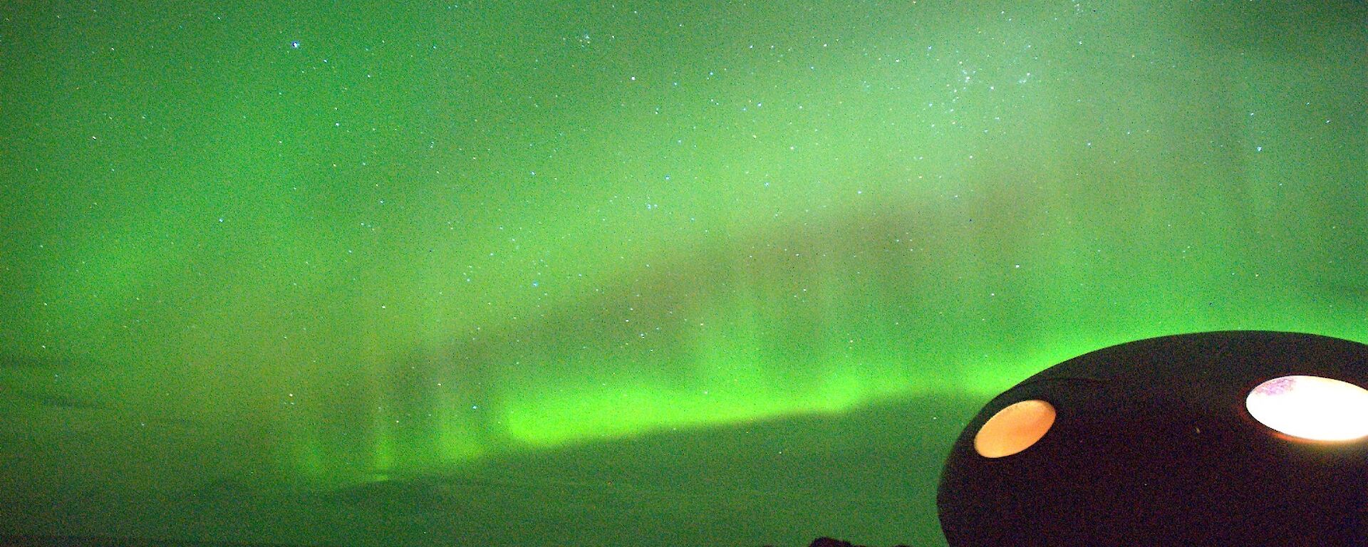 Fluorescent band of green lights running horizontally across the night sky behind an oval shaped field hut called a Googie